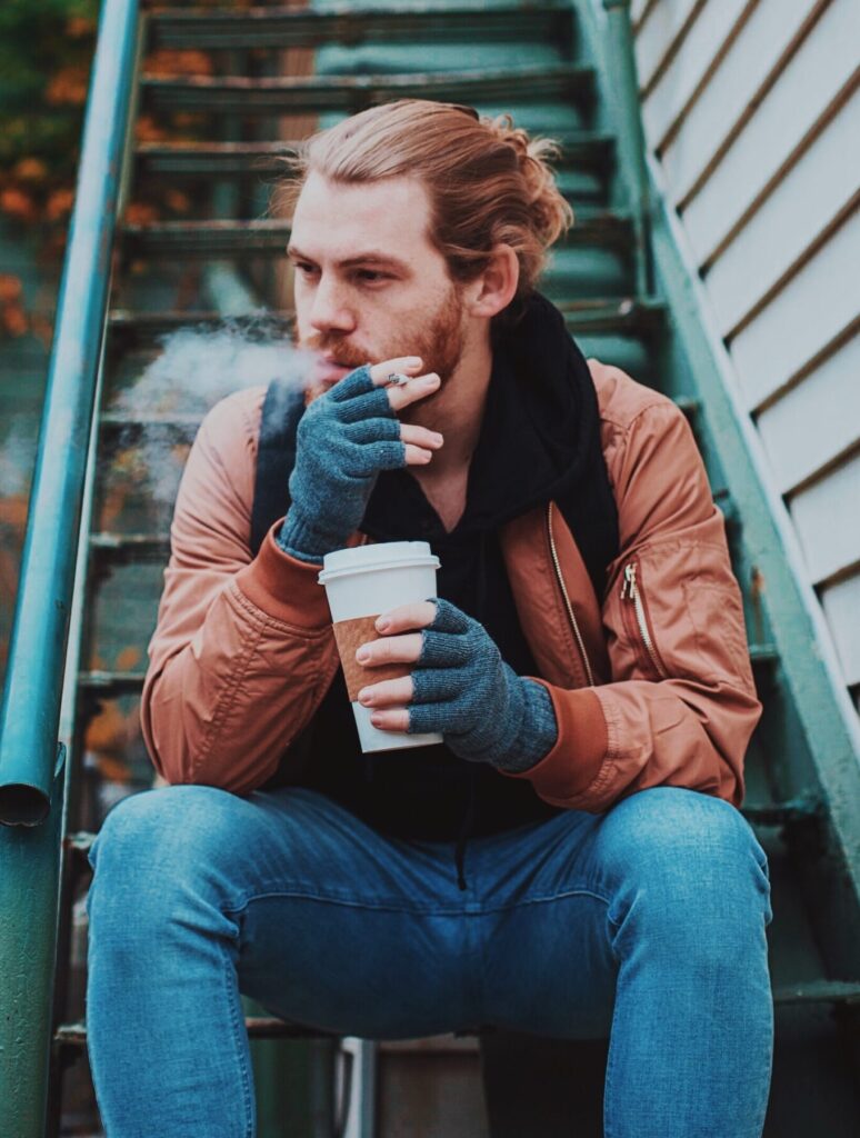 A young man sits on outdoor metal stairs, holding a coffee cup and smoking. He wears a brown jacket, blue jeans, gloves, and a scarf. There's visible vapor from his breath or smoke as he considers the power of hypnotherapy.