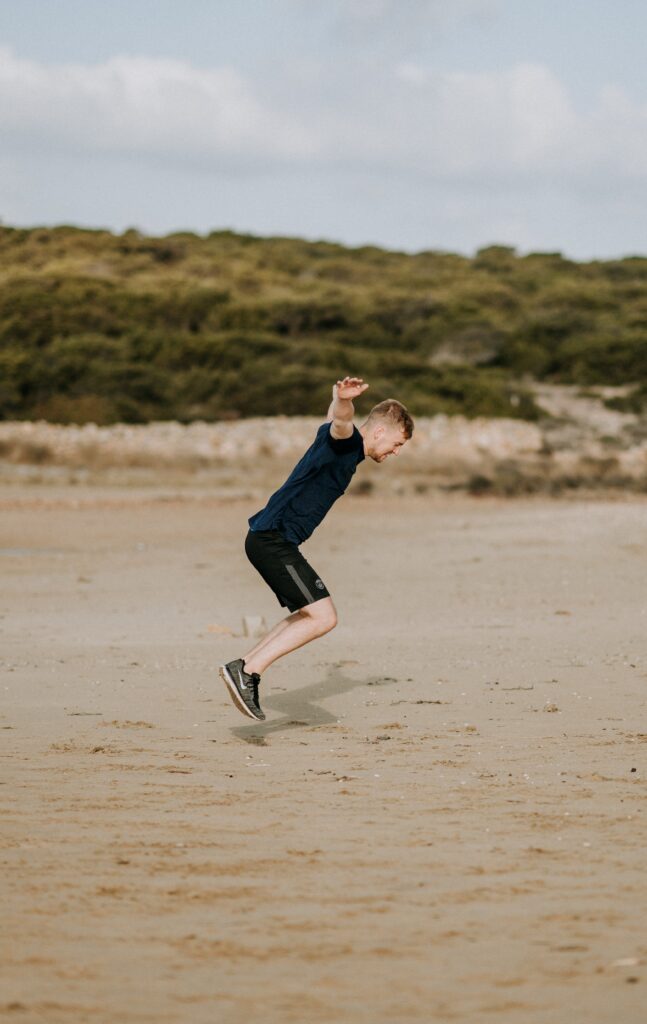 A young boy jumps energetically on a sandy beach, wearing a dark t-shirt and shorts, with a blurred natural landscape in the background. his expression is focused and full of joy.