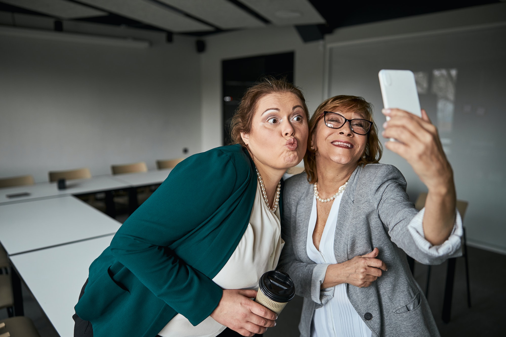 Two women in business attire taking a selfie in a conference room, one making a playful face while the other smiles, holding a coffee cup and smartphone.