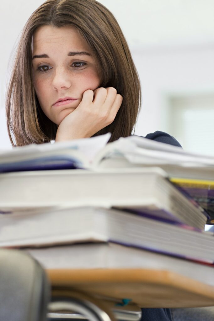 A young woman looking worried or overwhelmed, possibly experiencing school anxiety, resting her chin on her hand, with a stack of books in front of her, studying or doing research.
