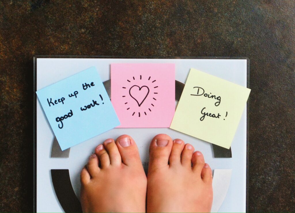 A person's bare feet standing on a weight management scale, with three colorful sticky notes on it; one blue with "Keep up the good work!", one pink with a heart, and one yellow saying "Doing great!".