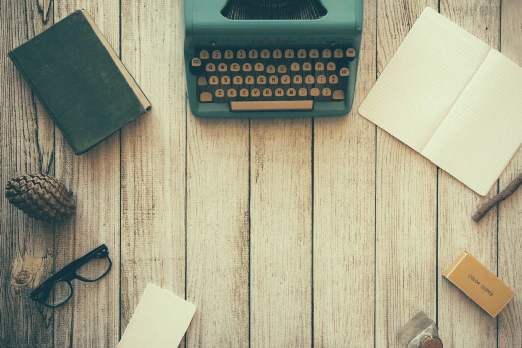 Vintage typewriter with a book, eyeglasses, notebook, pine cone, and sticks on a wooden table, suggesting a creative or writing setting.