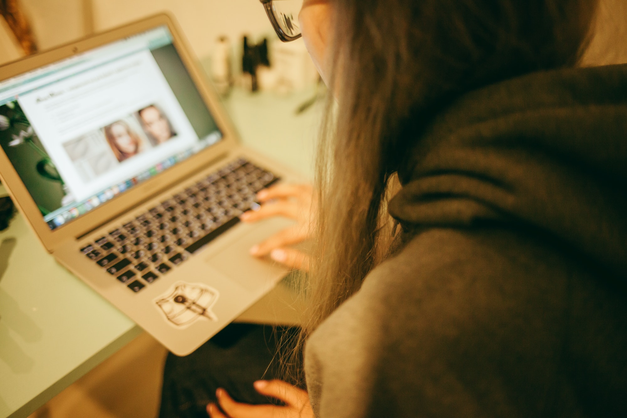 A person with long hair wearing glasses and a hoodie browses a website on a laptop, displaying profiles of people. The focus is on the laptop screen and the user's hands typing.