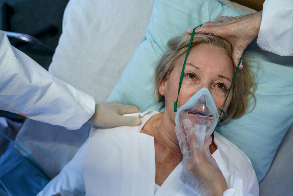 A female patient lying in a hospital bed wearing an oxygen mask, with a healthcare worker placing a hand on her forehead, showing concern and care about the potential benefits of hypnotherapy.