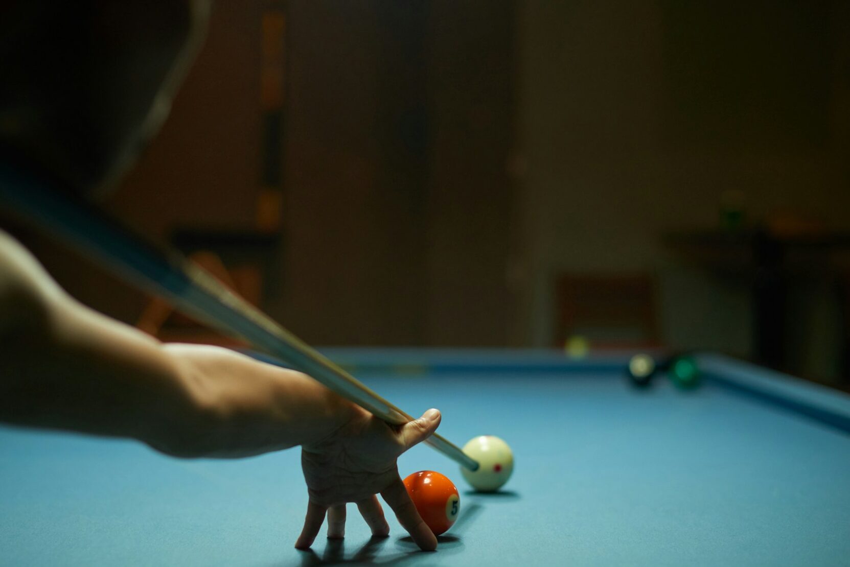 A person engaged in sports hypnotherapy, focusing on hitting the cue ball with a pool stick, aiming at an orange-striped ball on a blue felt table, with other balls and the shadowed room in the background.