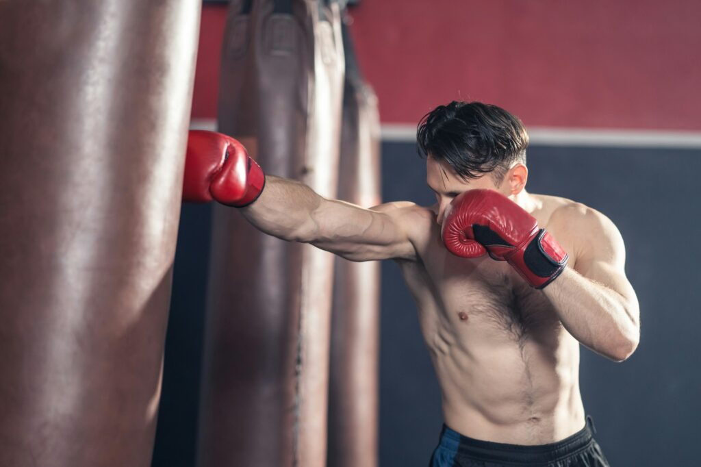 A muscular man wearing red boxing gloves is throwing a punch at a heavy bag in a gym. His focus and strength, enhanced by sports hypnotherapy, are evident as he makes contact with the bag.