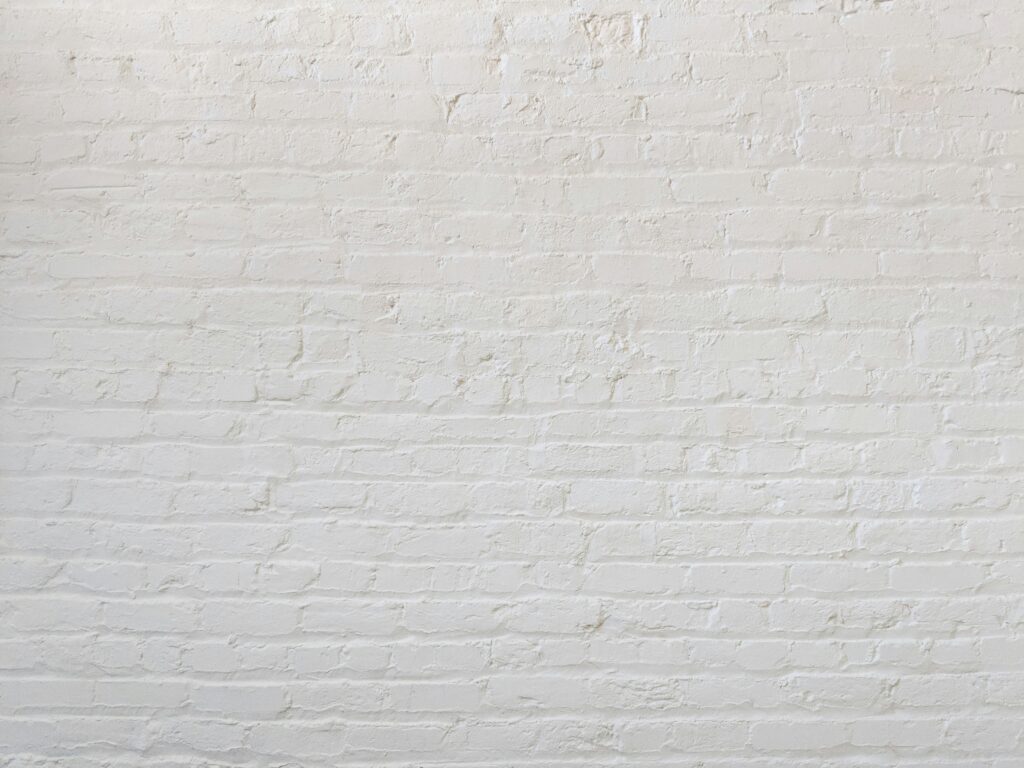 A white brick wall is shown, with slightly uneven texture and minor imperfections. The bricks and mortar exhibit a painted surface, providing a clean and simple backdrop. The lighting is even, highlighting the wall's details and texture.