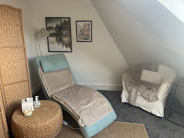 A cozy room invites tranquility, featuring a padded lounge chair with a tan covering, a white armchair with a cushion and throw, and a wicker ottoman. Wall art decorates the slanted wall while the power of hypnotherapy is subtly reflected through serene tones. A floor lamp casts gentle light on the small table holding a candle and bottle.