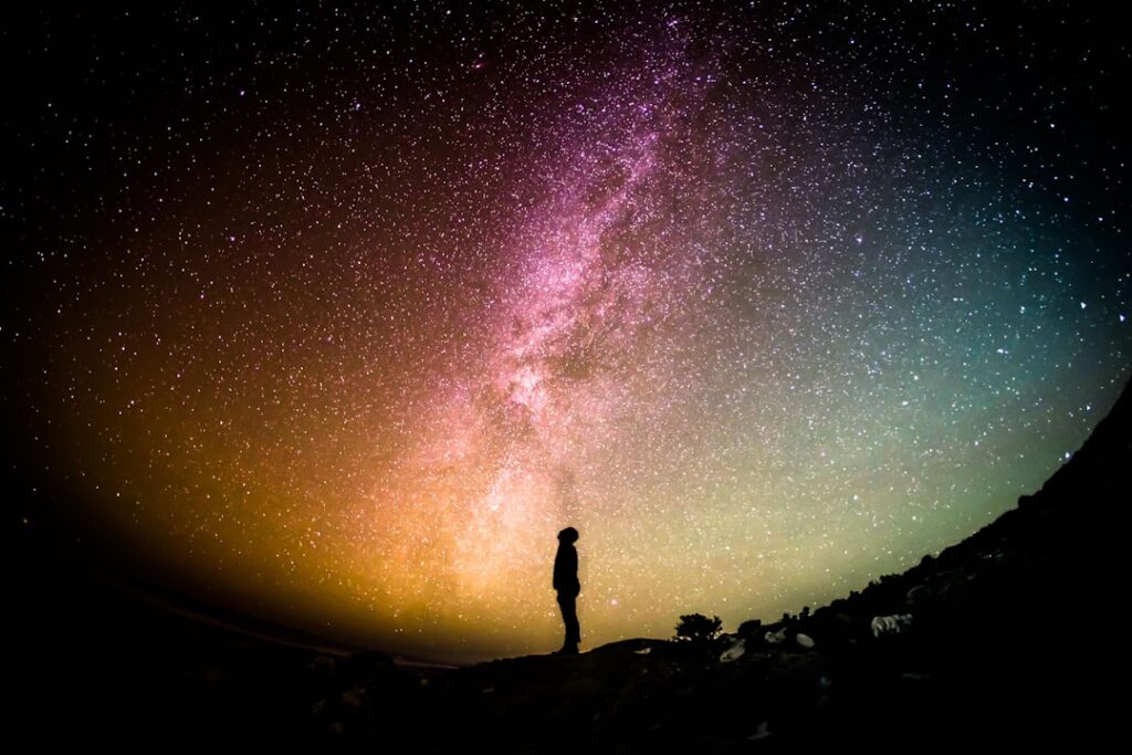 Silhouette of a person standing on a hill, gazing up at a colorful starry night sky featuring the Milky Way galaxy. The horizon glows with shades of orange, pink, and green, creating a vibrant celestial panorama.