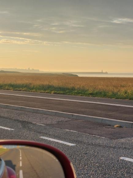 A coastal landscape with a grassy field, a road in the foreground, and an offshore oil platform in the distance. The image is taken from a vehicle, with part of a red side mirror visible on the left.