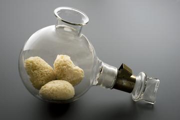A round glass chemical flask lies on its side, containing three yellowish, sponge-like objects inside, sealed with a metal and glass stopper—evoking the spirit of modern healing and hypnotherapy—resting on a smooth gray surface.